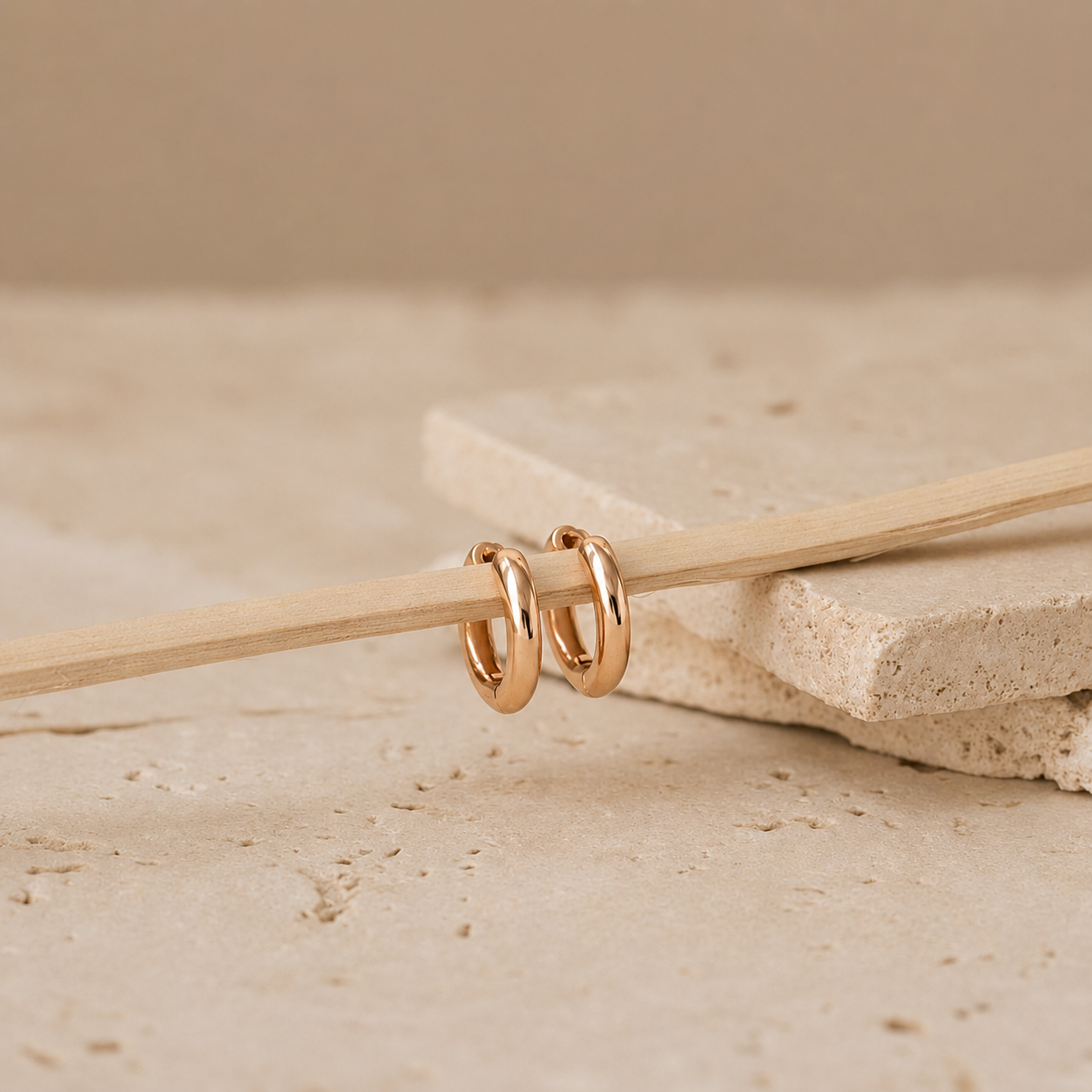 A pair of small gold hoop earrings resting on a thin wooden stick, placed on stacked stone slabs against a warm beige background.